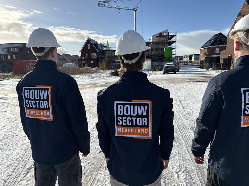 Drie vakmensen van Bouwsector Nederland met witte helm en donkere jas lopen door een nieuwbouwstraat met een bouwkraan